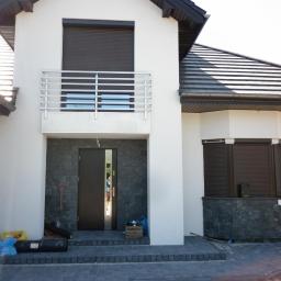 ADD Projekt - Nowoczesny dom jednorodzinny w Tarnobrzegu z zamontowanymi zewnętrznymi roletami w kolorze ciemnego brązu i otwartymi, ciemnymi drzwiami wejściowymi. Widoczny balkon z metalową balustradą.