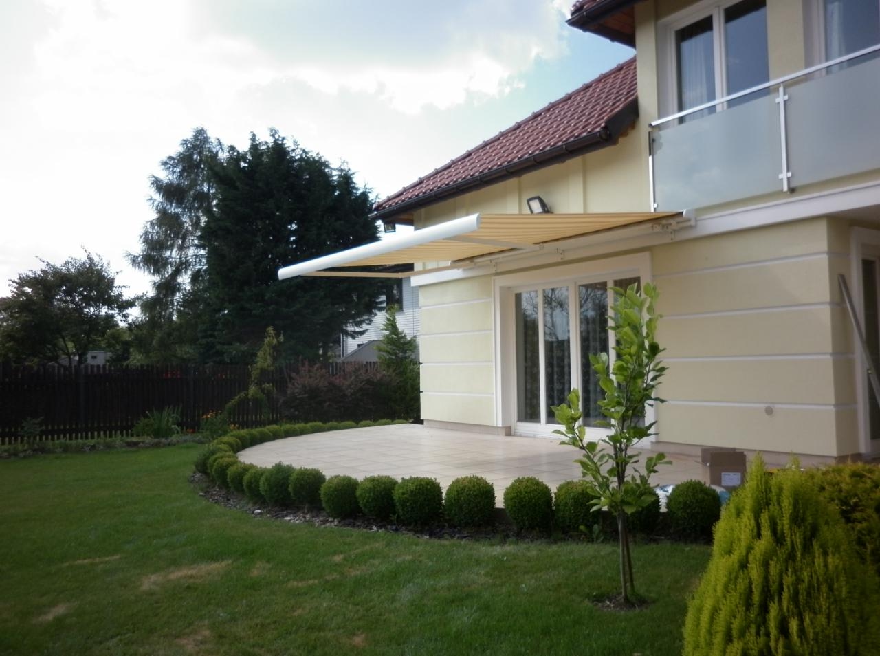 Żółta markiza tarasowa zamontowana nad przesuwnymi drzwiami patio, z widokiem na zadbany trawnik i dekoracyjne krzewy.