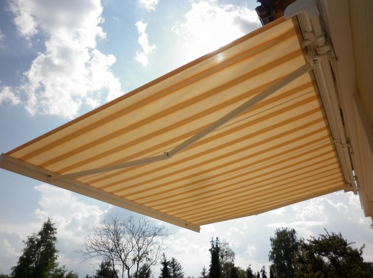 Rozwinięta markiza w paski koloru beżowo-żółtego, zamontowana na elewacji budynku, z widokiem na błękitne niebo z chmurami i korony drzew