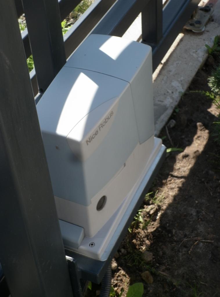 Szary automat do bram przesuwnych Nice Robus zamontowany przy słupku ogrodzenia, widoczne okablowanie i podłoże z ziemią i roślinnością.