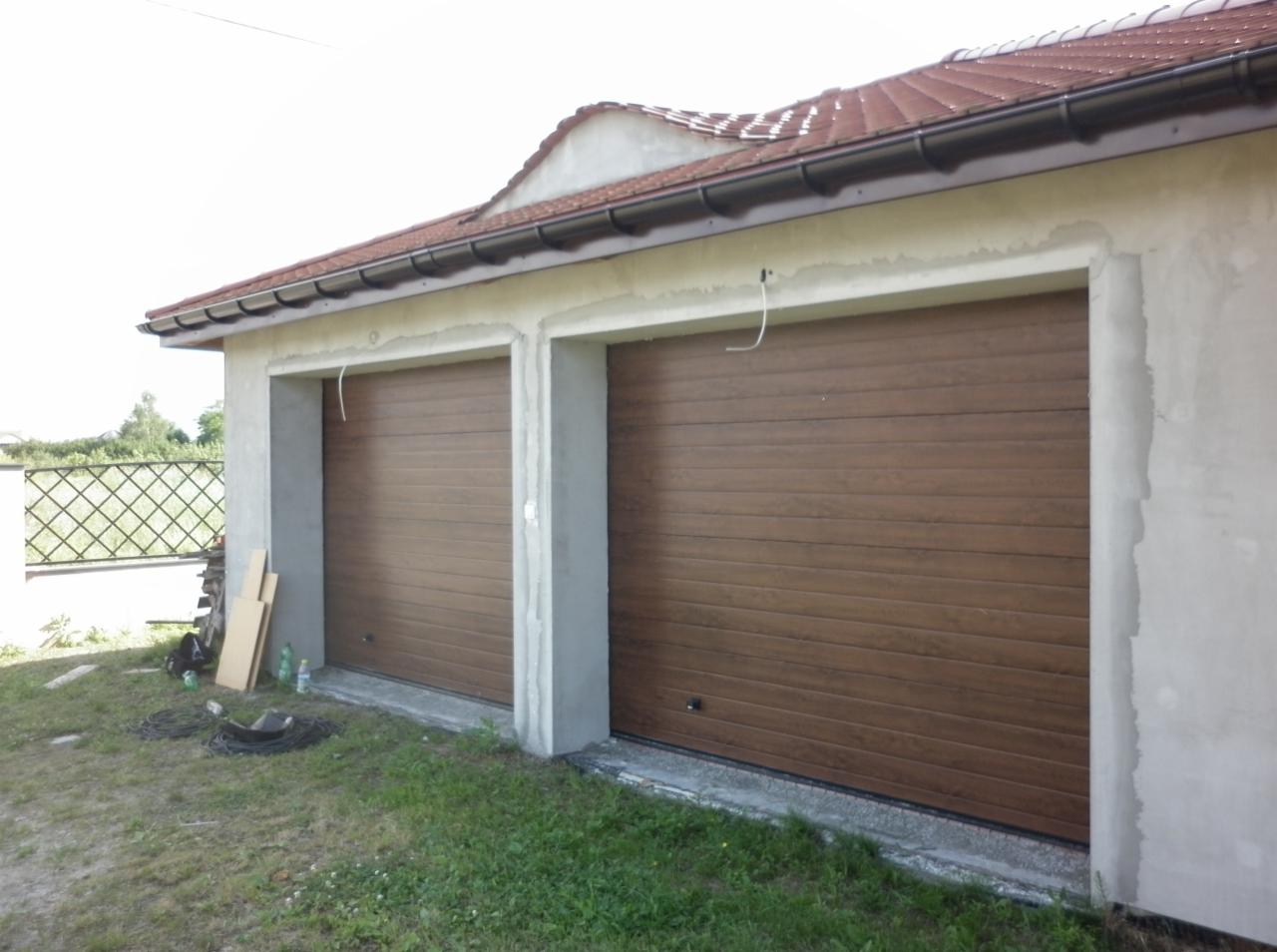 Dwa brązowe segmentowe bramy garażowe w surowym stanie budynku z widocznymi przewodami elektrycznymi, materiałami budowlanymi i trawą.