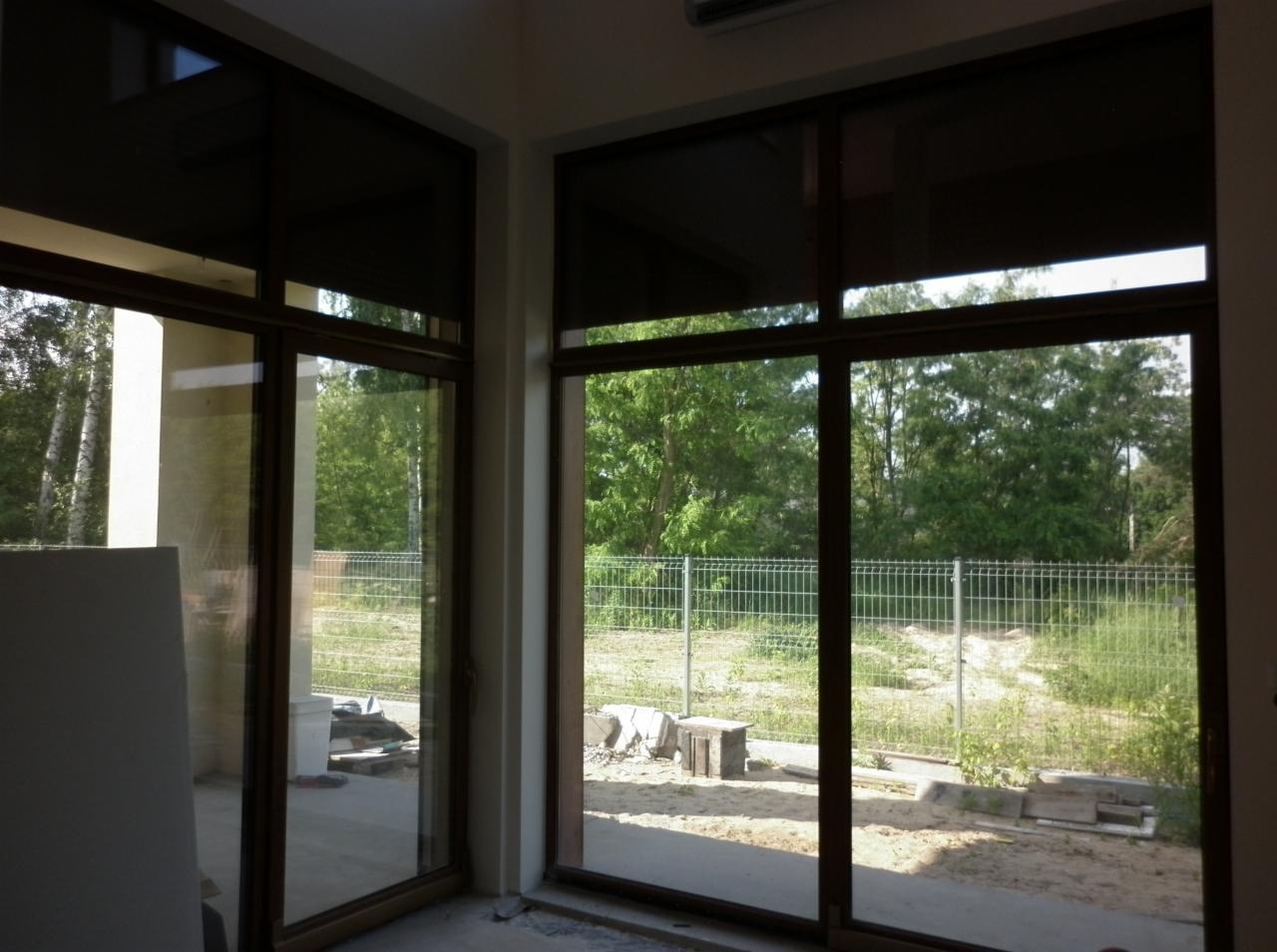 Widok z wnętrza domu przez duże, przesuwne okna z brązowymi ramami i opuszczonymi zasłonami; za oknami widoczny ogród z siatką ogrodzeniową.