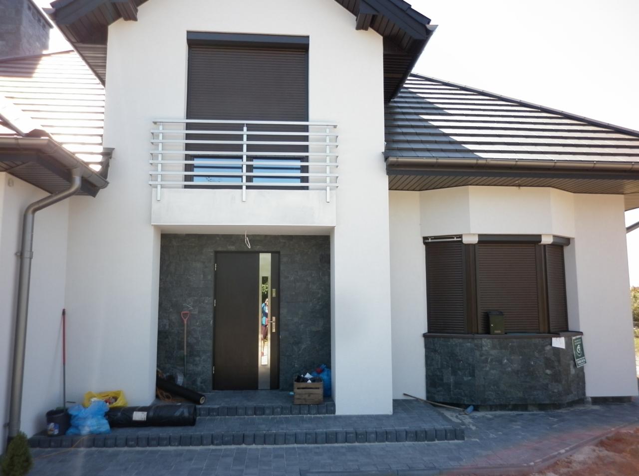 Nowoczesny dom jednorodzinny w Tarnobrzegu z zamontowanymi zewnętrznymi roletami w kolorze ciemnego brązu i otwartymi, ciemnymi drzwiami wejściowymi. Widoczny balkon z metalową balustradą.