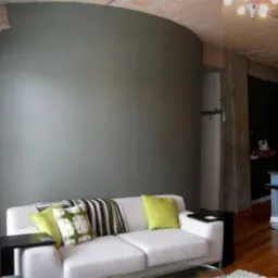 Jasny salon z szarą, zaokrągloną ścianą i białą sofą z zielonymi poduszkami. Widoczny fragment kuchni i drewniana podłoga. Loftowy styl wnętrza.