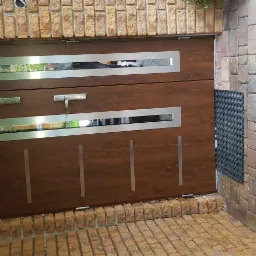 Brązowa brama garażowa segmentowa z poziomymi przeszkleniami w srebrnych ramach, zamontowana w murze z cegły klinkierowej. Na posadzce z kostki brukowej leżą sportowe buty.