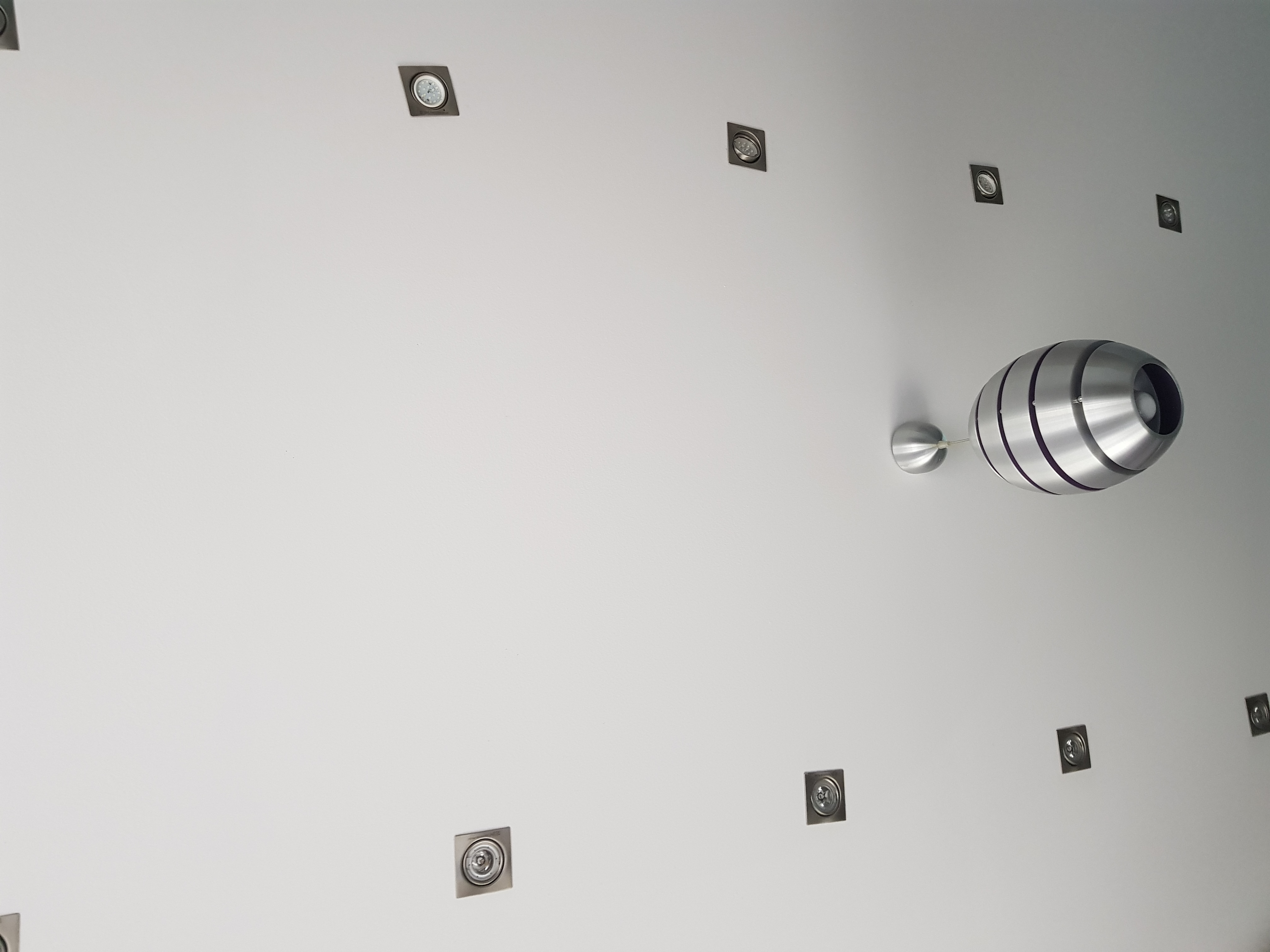 Biały sufit z zamontowanymi kwadratowymi lampami punktowymi i nowoczesną, metalową lampą wiszącą w kształcie torpedy.