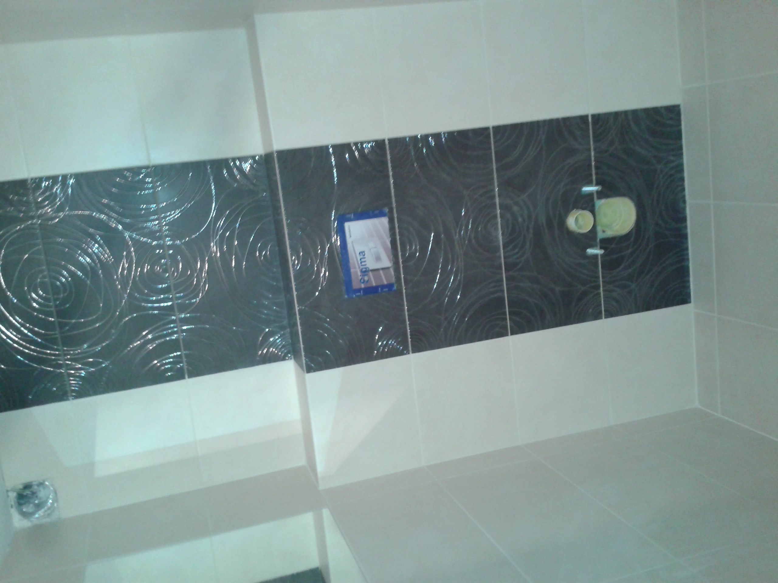 Wykończona łazienka z ułożonymi białymi i czarnymi płytkami z motywem spirali, widoczne elementy instalacji sanitarnej i naklejka producenta na jednej z płytek.