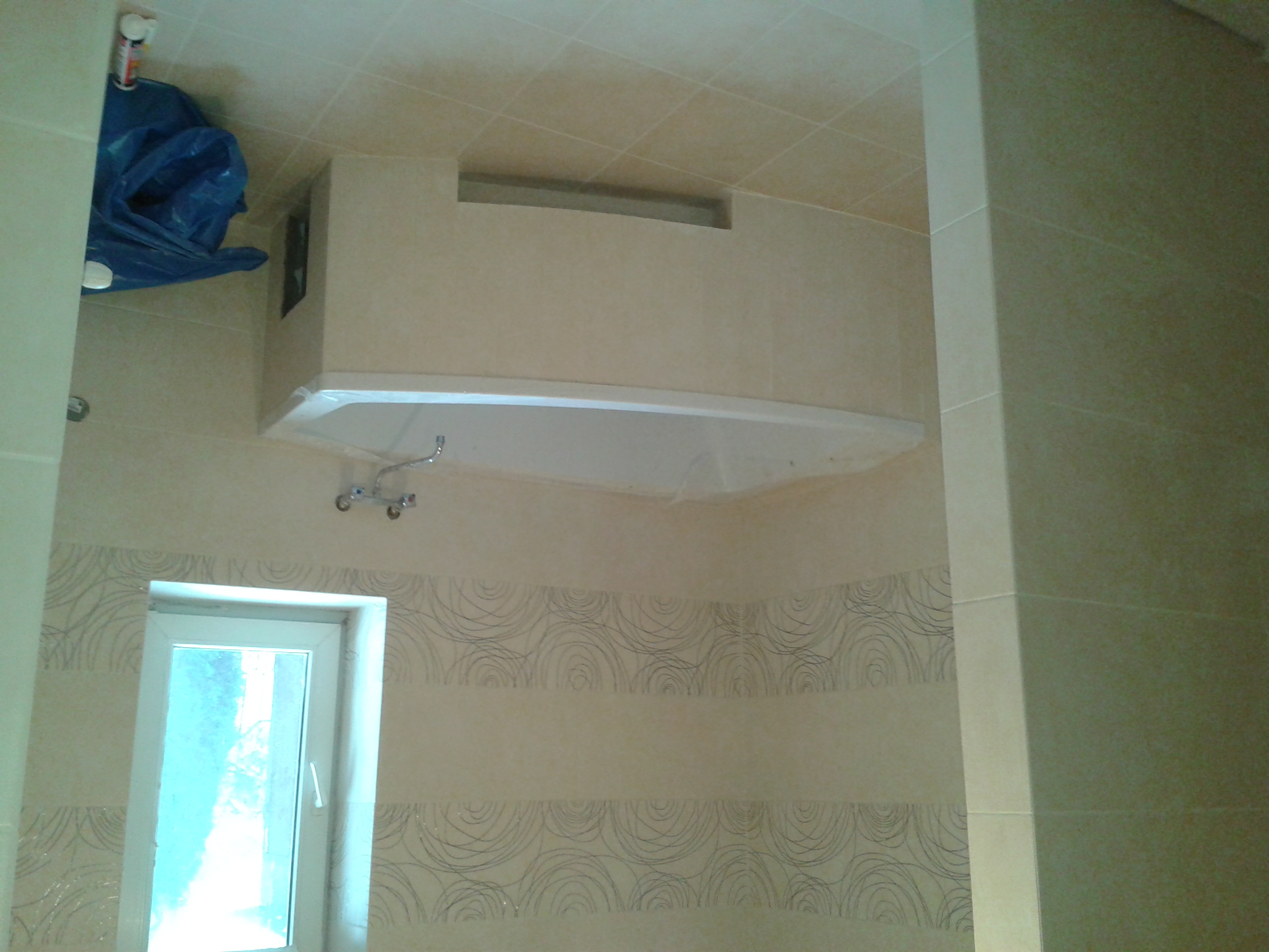 Biała wanna zabudowana w jasnej łazience w Wesołej k. Dynowa, widoczne płytki z delikatnym wzorem, okno i zabezpieczona niebieską folią armatura.