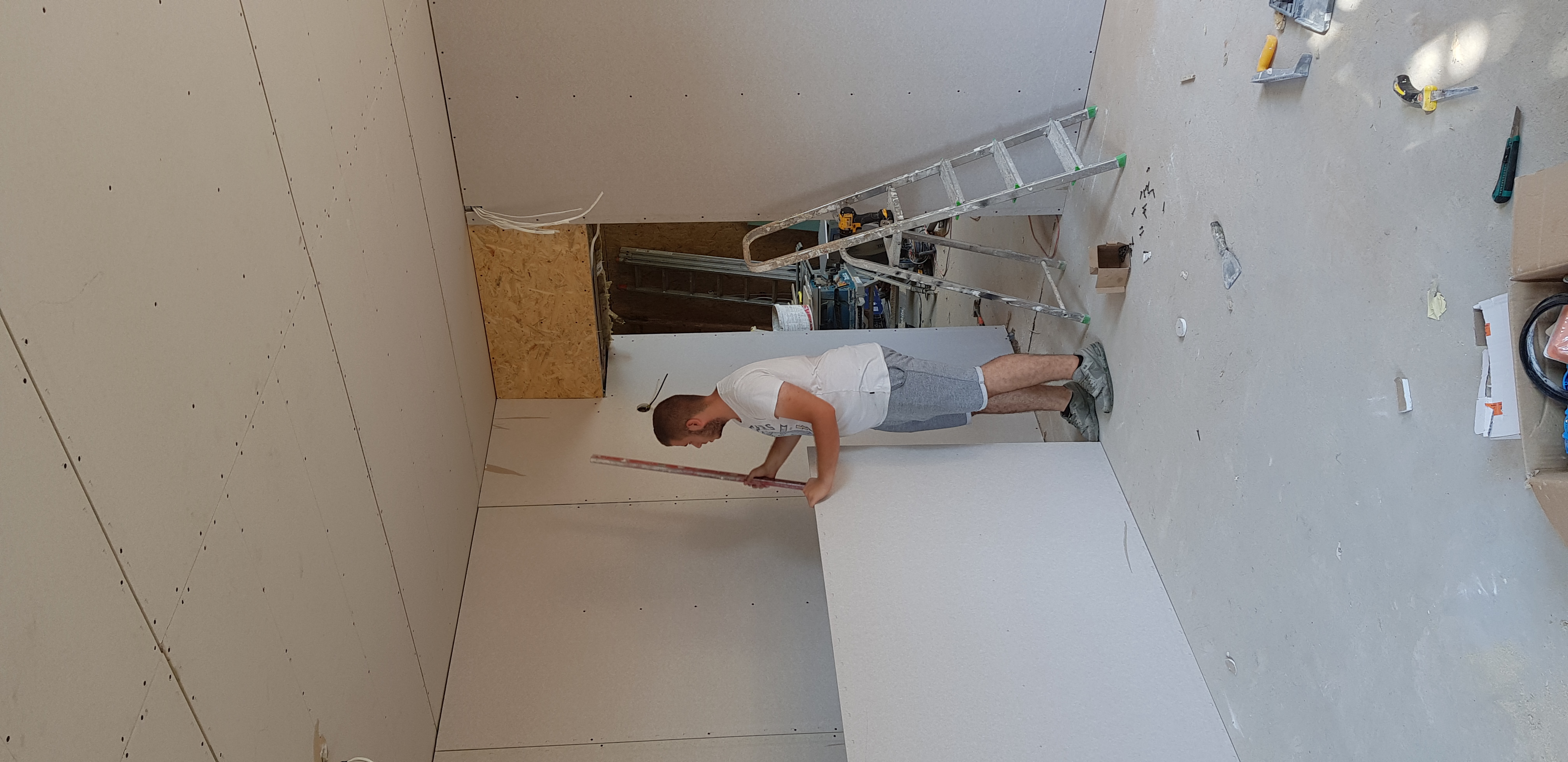 Mężczyzna w szarej koszulce i spodenkach, przy użyciu poziomicy, sprawdza pionowość nowo zamontowanej płyty gipsowo-kartonowej w surowym wnętrzu. W tle widoczna drabina, narzędzia i fragment...