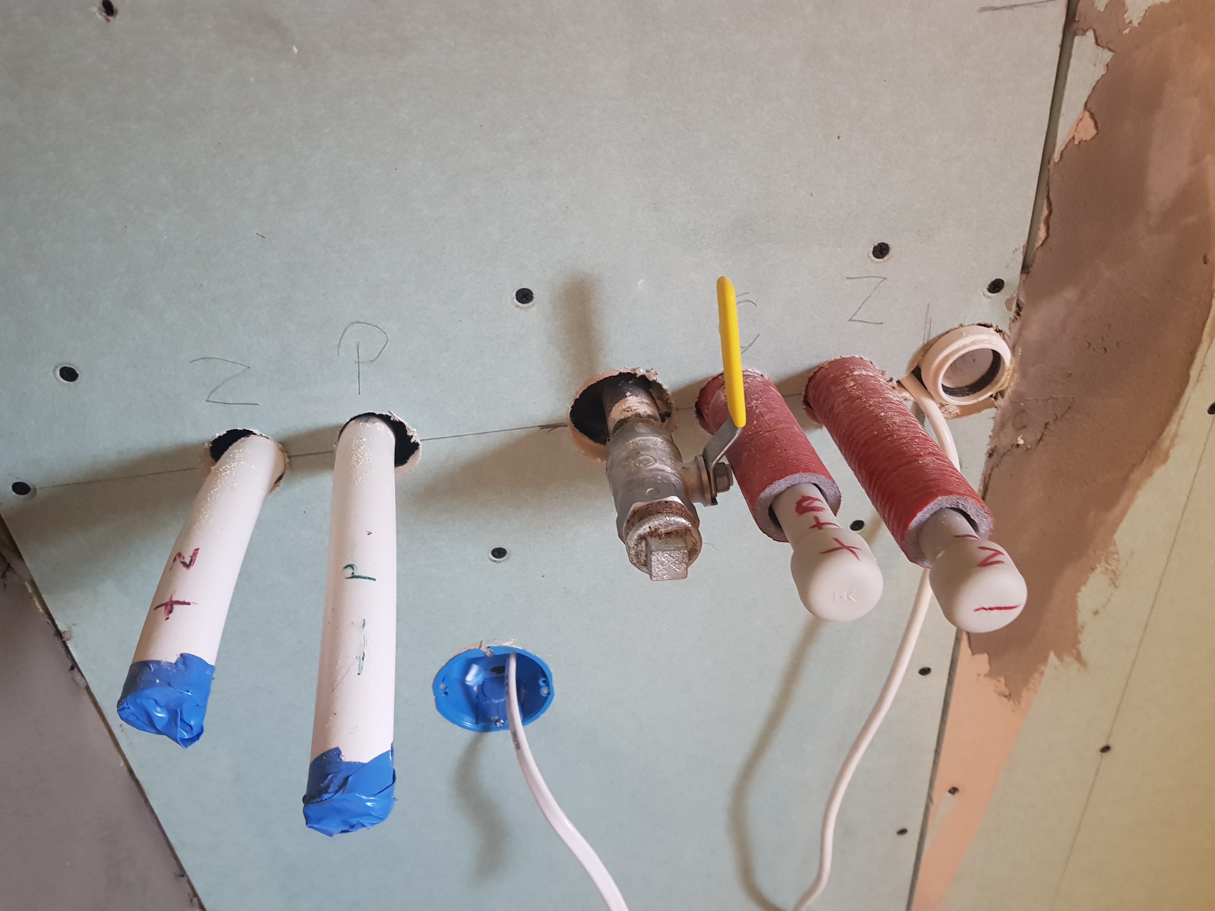 Ściana z płyt kartonowo-gipsowych z wystającymi rurami instalacji wodnej i elektrycznej, oznaczone literami 'Z' i 'P', z widocznymi śladami po wierceniu i niewielkimi uszkodzeniami na krawędziach.