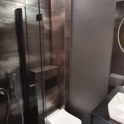 P.H.U. Filip Dutkiewicz - Nowoczesna łazienka z ciemnymi płytkami imitującymi metal, kabiną prysznicową typu walk-in, podwieszaną toaletą i szafką umywalkową z ryflowanym frontem, oświetlona ledowym pasem nad sufitem.