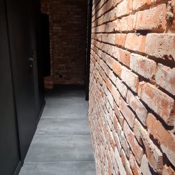 P.H.U. Filip Dutkiewicz - Wąski korytarz z ceglaną ścianą, szarą podłogą z płytek i czarnymi, punktowymi lampami sufitowymi.