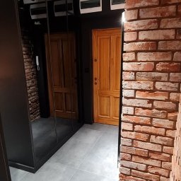 P.H.U. Filip Dutkiewicz - Nowoczesny przedpokój z ceglaną ścianą, szarą podłogą, czarną szafą z lustrami i drewnianymi drzwiami, oświetlony reflektorami na szynie.