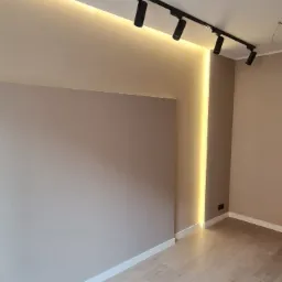 Nowoczesne oświetlenie sufitowe z czarnymi reflektorami na szynie, podświetlenie LED w pionowej wnęce ściany, jasne panele podłogowe, beżowe ściany.