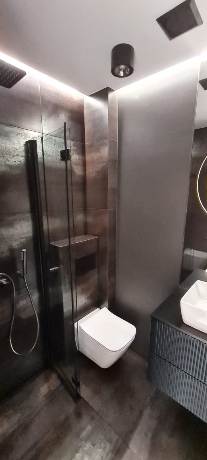 Nowoczesna łazienka z ciemnymi płytkami imitującymi metal, kabiną prysznicową typu walk-in, podwieszaną toaletą i szafką umywalkową z ryflowanym frontem, oświetlona ledowym pasem nad sufitem.