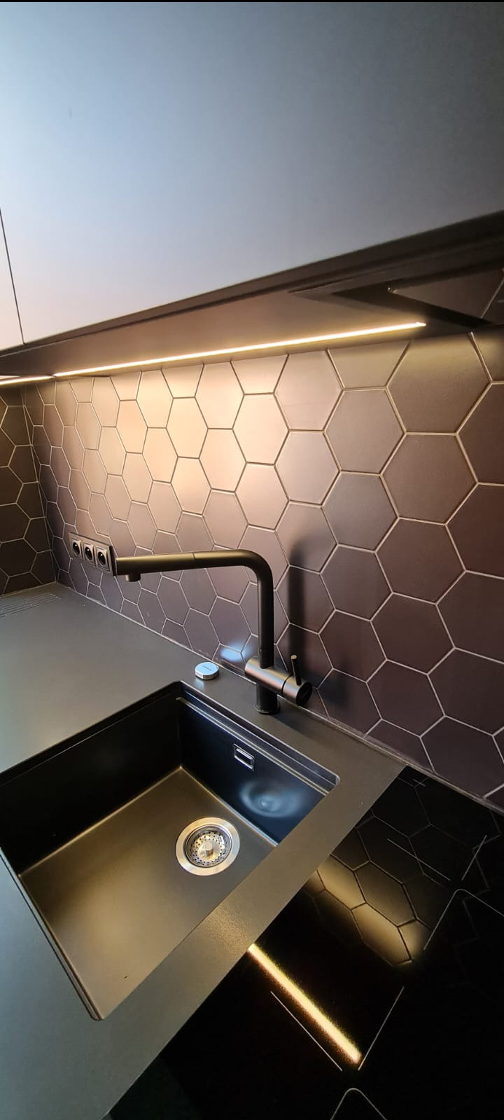 Czarny zlewozmywak wkomponowany w czarny blat kuchenny, z baterią w tym samym kolorze, na tle ściany z heksagonalnych płytek w odcieniach szarości i brązu, oświetlonych listwą LED.
