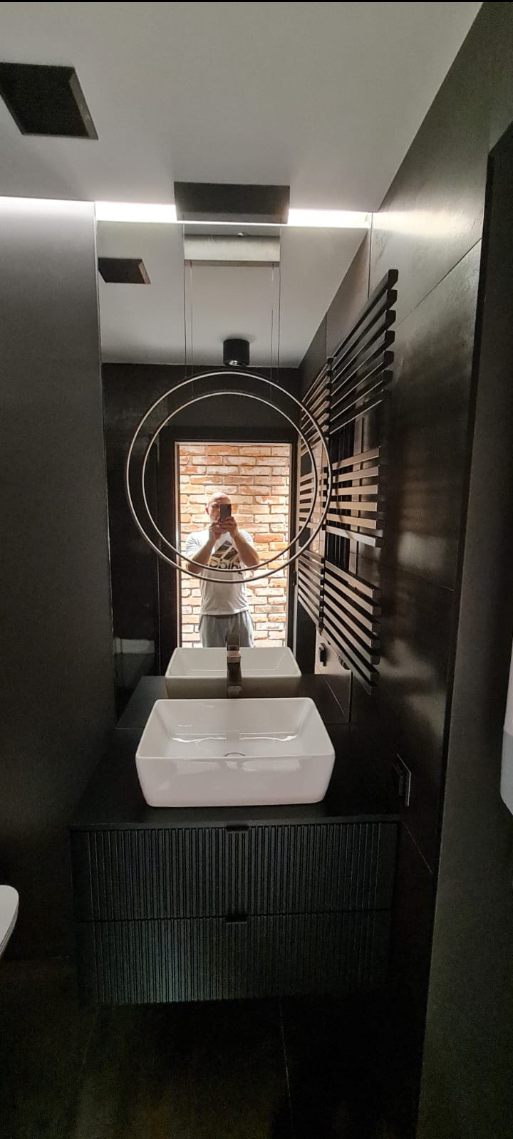 Nowoczesna łazienka z czarnymi ścianami, białą umywalką nablatową i okrągłą lampą wiszącą, w lustrze odbija się ceglana ściana i osoba robiąca zdjęcie.