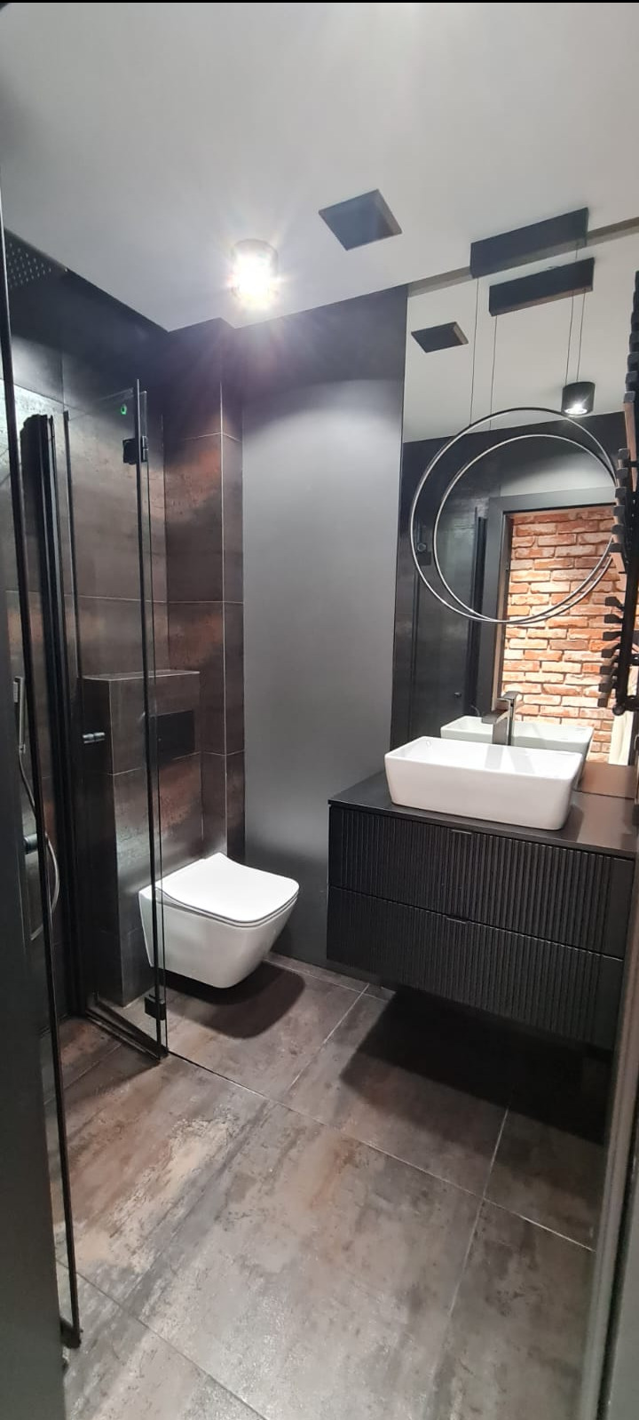 Nowoczesna łazienka z kabiną prysznicową, podwieszaną toaletą, umywalką nablatową na czarnej szafce z frezowanymi frontami, okrągłym lustrem i płytkami imitującymi rdzę na ścianach i podłodze.