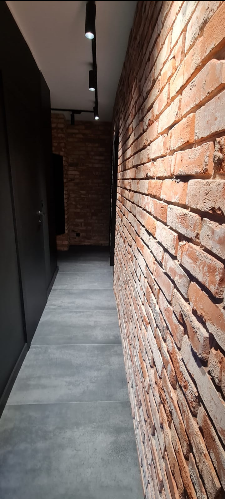 Wąski korytarz z ceglaną ścianą, szarą podłogą z płytek i czarnymi, punktowymi lampami sufitowymi.