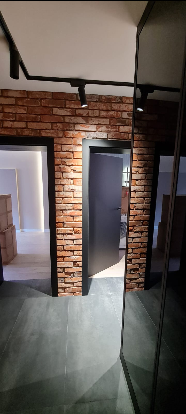 Wnętrze po remoncie: ściana z cegły klinkierowej, dwie czarne ościeżnice z szarymi drzwiami, szara podłoga z płytek, czarne reflektory na suficie.