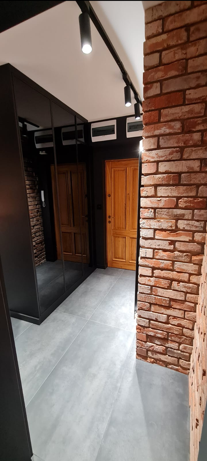 Nowoczesny przedpokój z ceglaną ścianą, szarą podłogą, czarną szafą z lustrami i drewnianymi drzwiami, oświetlony reflektorami na szynie.