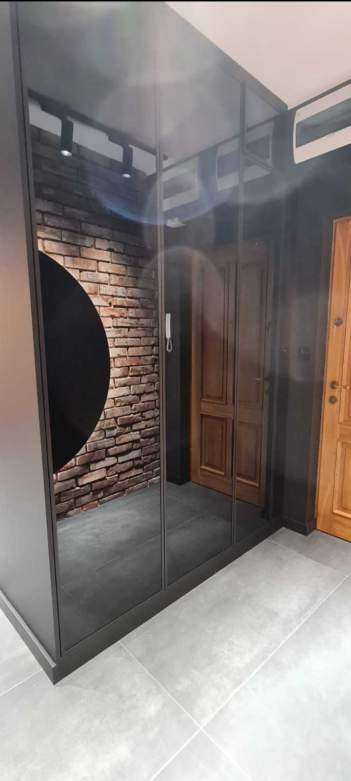 Nowoczesna szafa wnękowa z lustrzanymi drzwiami przesuwnymi, odbijająca ceglaną ścianę z dekoracyjnym czarnym okręgiem i drewniane drzwi wejściowe, szara podłoga z płytek.
