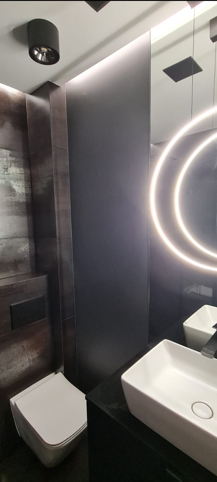 Nowoczesna łazienka w odcieniach szarości i czerni z podświetlanym lustrem, wiszącą toaletą i prostokątną umywalką nablatową.