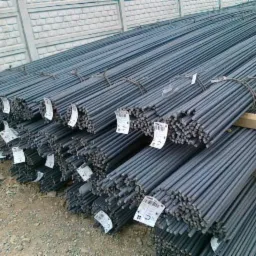 Sztaple wiązek prętów zbrojeniowych z etykietami, ułożone na ziemi przy betonowym ogrodzeniu.