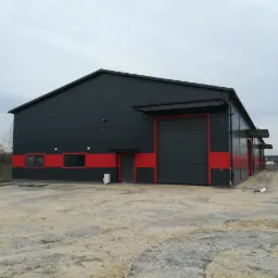 Zewnętrzny widok nowo wybudowanej hali stalowej w kolorze grafitowym z czerwonym pasem, z widocznymi drzwiami wejściowymi i bramą garażową, na placu budowy z kostką brukową i ziemią.