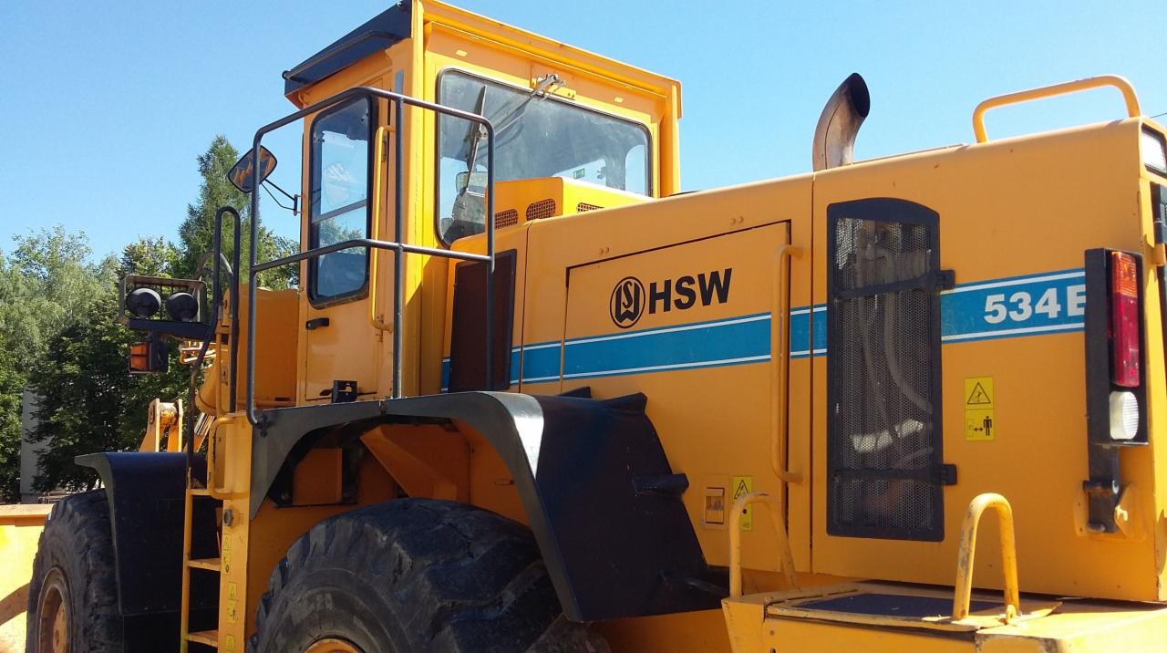 Żółta ładowarka HSW 534 E z widocznym logo firmy i oznaczeniami bezpieczeństwa, sfotografowana z boku na tle błękitnego nieba i zieleni drzew.