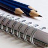 Spiralny notatnik z dwoma ołówkami na białym tle. Ołówki częściowo poza kadrem. Przybliżenie na detale notatnika i grafitu ołówków.