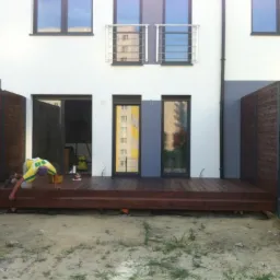 Montaż drewnianego tarasu przy nowoczesnym domu. Pracownik w żółtej koszulce nanosi impregnat na deski. Widoczne narzędzia i ogrodzenie.