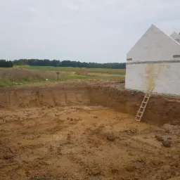 Wykop pod fundamenty domu jednorodzinnego w trakcie realizacji, widoczna drewniana drabina oparta o ścianę wykopu, w tle surowy budynek z białego bloczka.