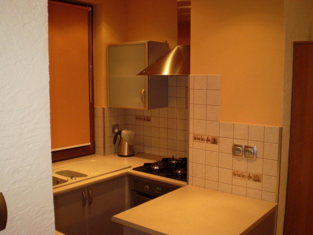 Umeblowana kuchnia z beżowymi płytkami i szafkami. Widoczny okap, zlew i okno z pomarańczową roletą. Na płytkach dekoracyjne napisy.