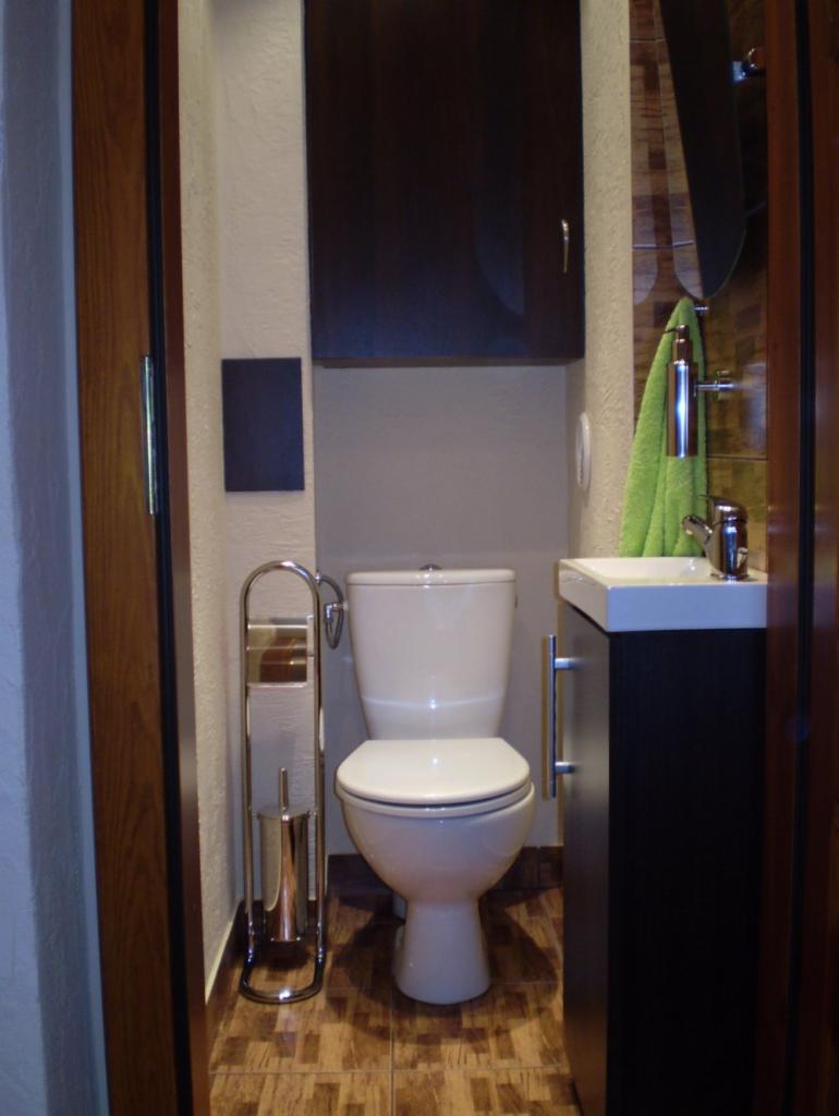 Wnętrze wyremontowanej, małej łazienki z toaletą, szafką pod umywalką i akcesoriami. Na ścianie lustro i szafka. Brązowe płytki na podłodze.