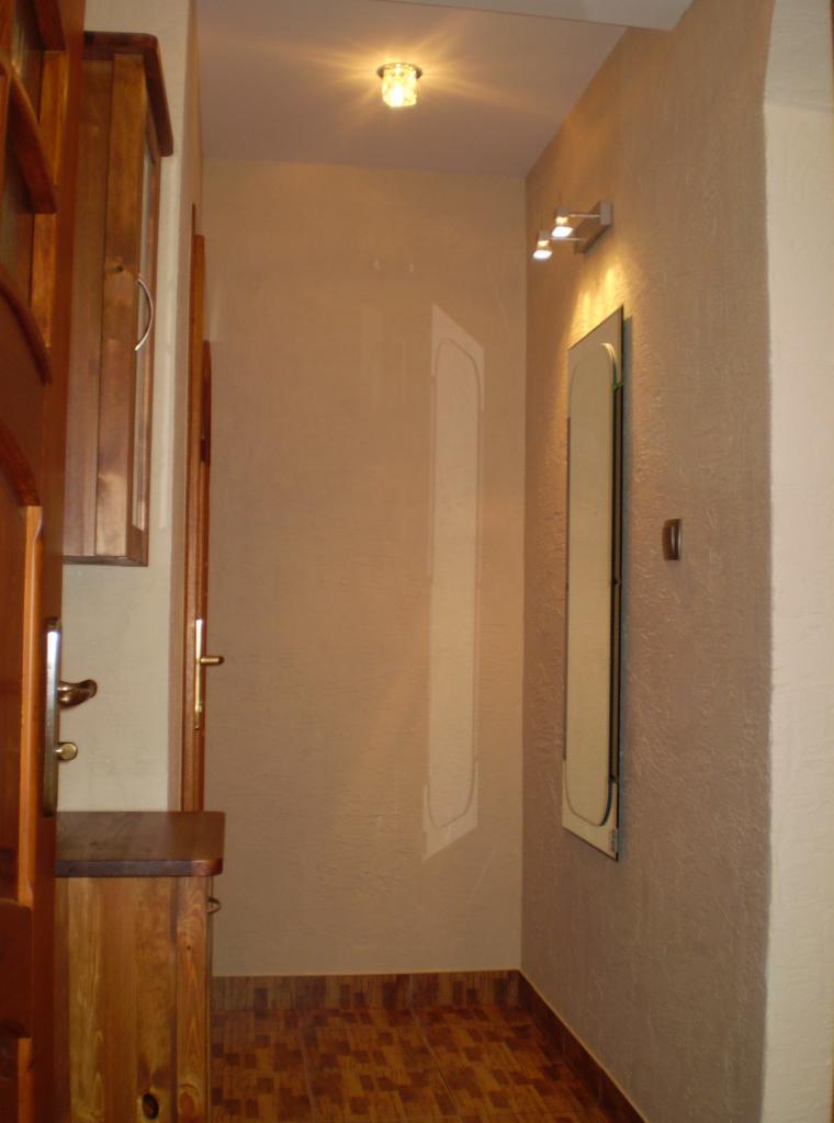 Beżowy korytarz z drewnianą podłogą, szafką i lustrem. Na ścianie kinkiet z trzema punktami świetlnymi. Widoczna faktura ściany i oświetlenie sufitowe.