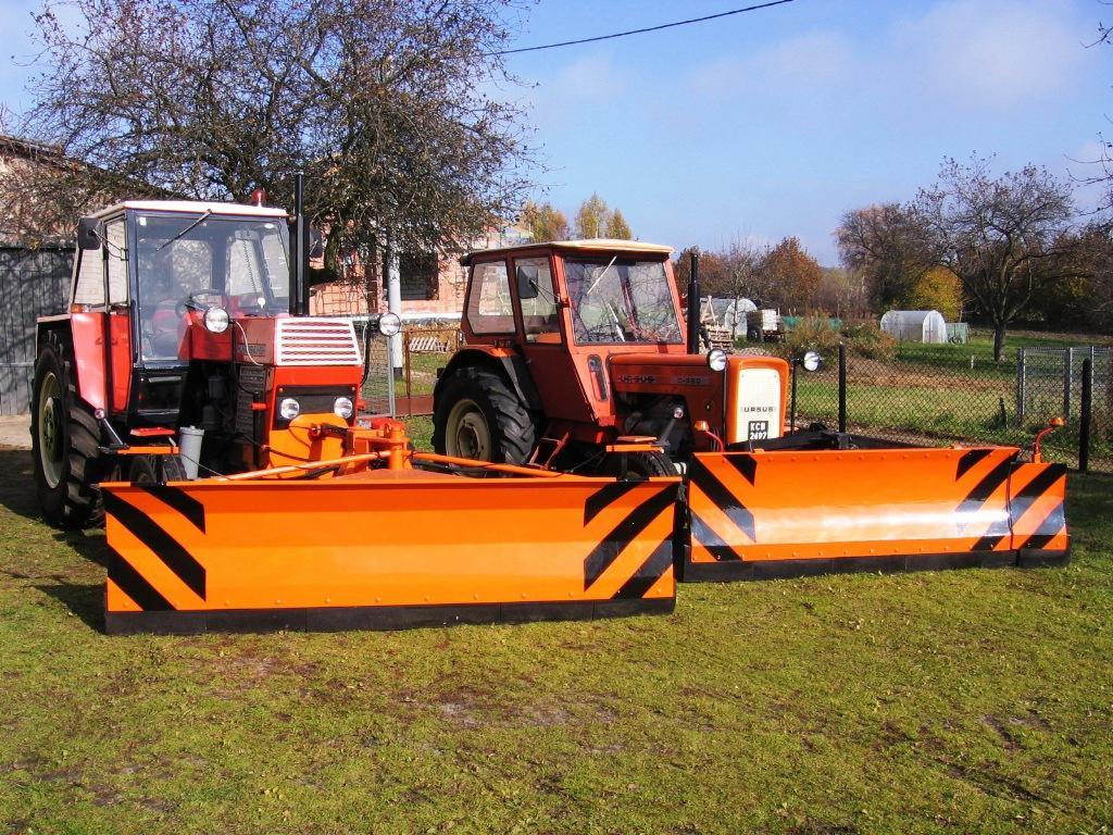 Dwa traktory Ursus z pomarańczowymi pługami na trawie. Jeden z białym dachem, drugi z pomarańczowym. W tle budynki i drzewa pod błękitnym niebem.