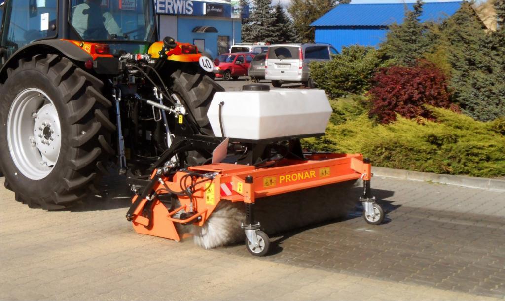 Traktor z pomarańczową szczotką Pronar do czyszczenia kostki brukowej na zewnątrz. W tle budynki i zieleń.