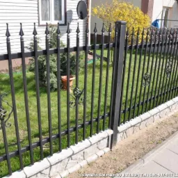 Nowoczesne, czarne ogrodzenie metalowe z ozdobnymi elementami, osadzone na kamiennej podmurówce, widoczne na tle zielonego trawnika i domu.