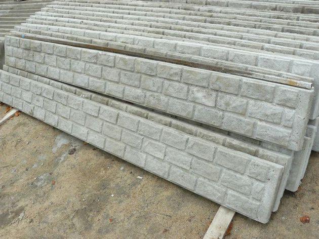 Stos szarych, betonowych prefabrykatów z fakturą cegły, ułożonych warstwami na ziemi. Elementy do budowy ogrodzeń lub ścian, widoczne detale tekstury betonu.