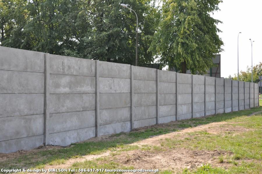 Długa, betonowa ściana działowa z widocznymi podziałami na segmenty, otoczona zielenią i drzewami w tle. Widok z perspektywy.