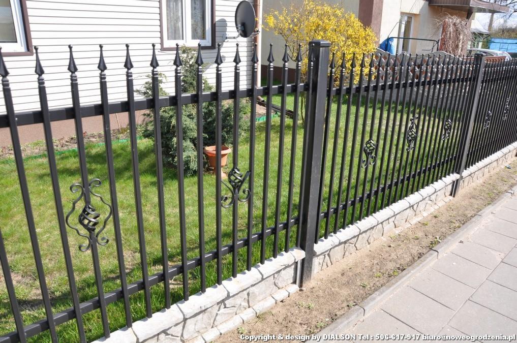 Nowoczesne, czarne ogrodzenie metalowe z ozdobnymi elementami, osadzone na kamiennej podmurówce, widoczne na tle zielonego trawnika i domu.