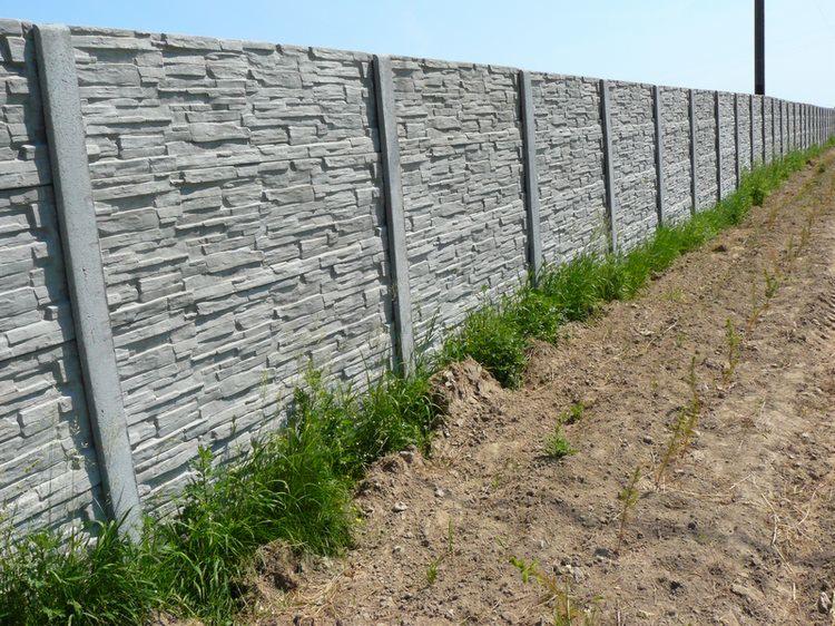 Długi, betonowy płot z fakturą kamienia, oddzielający teren porośnięty trawą od brązowej, ubitej ziemi. Widok z boku, w słoneczny dzień.