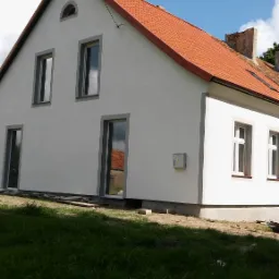 Boczny widok odnowionego domu z nową, czerwoną dachówką i świeżo pomalowaną, białą elewacją, na tle błękitnego nieba z chmurami, z widoczną skrzynką elektryczną na ścianie.