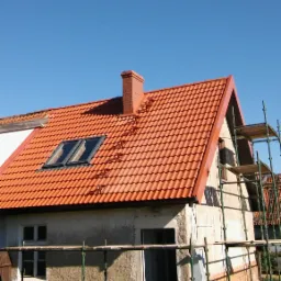 Dom w trakcie wymiany pokrycia dachowego na czerwoną dachówkę, widoczne rusztowania i okna dachowe.