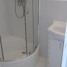 Biała narożna kabina prysznicowa z chromowaną armaturą i biała umywalka z szafką w czystej, jasnej łazience, widok zza uchylonych drzwi.