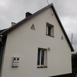 Elewacja domu jednorodzinnego po ociepleniu i pomalowaniu na jasny kolor, z brązowym dachem i elementami wykończeniowymi, widoczne okna i antena.