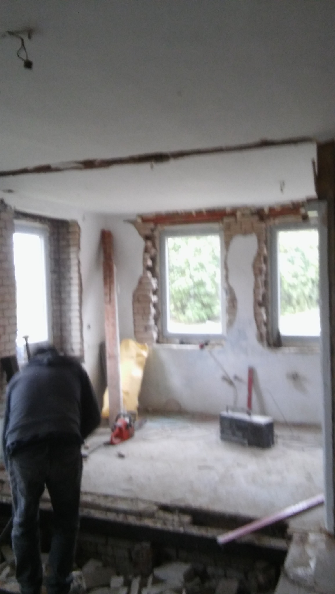 Demontaż ściany z cegły w trakcie przebudowy mieszkania, widoczne odsłonięte cegły wokół okien, narzędzia i pracownik w trakcie pracy.