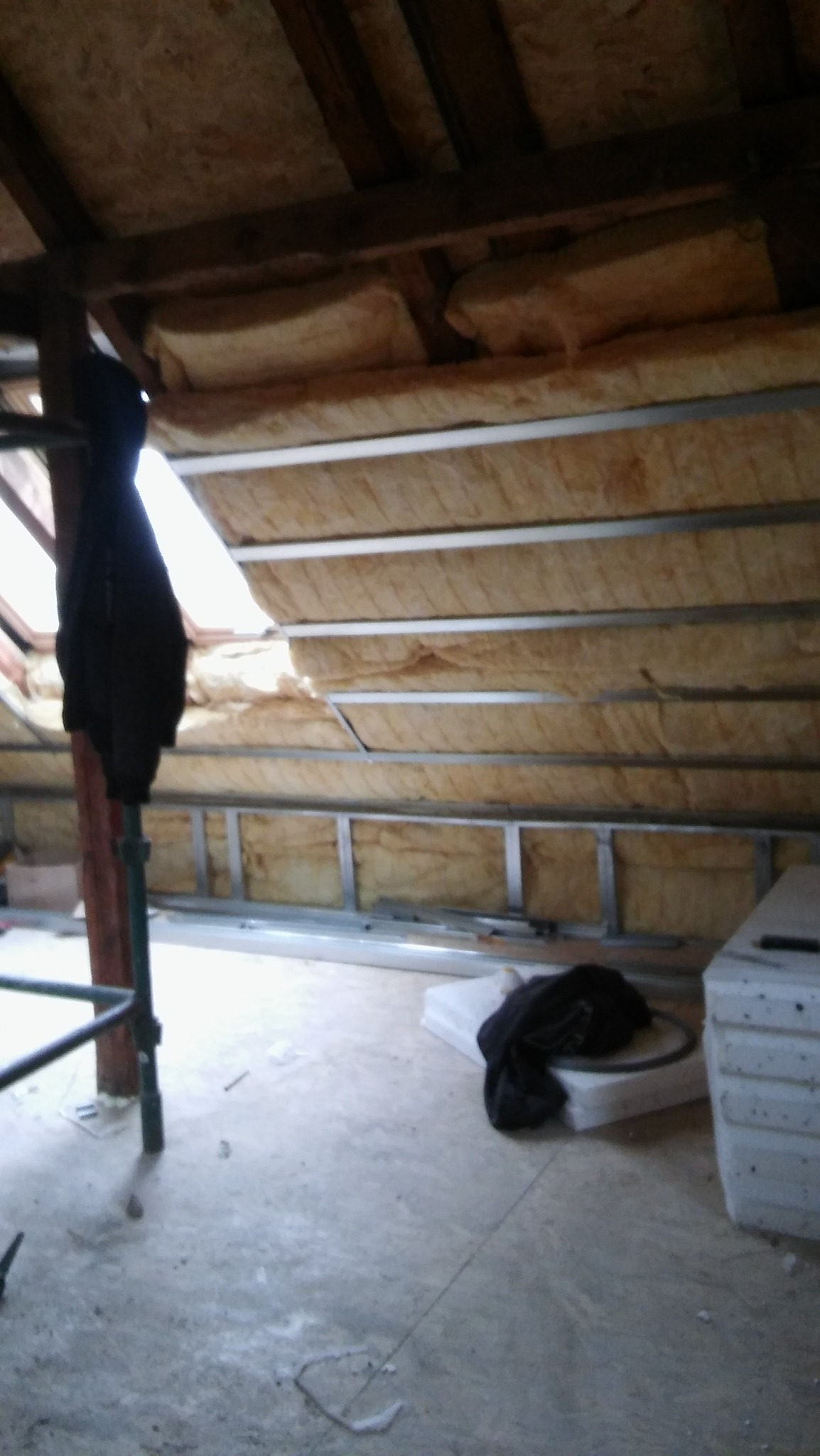 Poddasze w trakcie izolacji termicznej, widoczna wełna mineralna między metalowymi profilami, fragment okna dachowego i prowizoryczne rusztowanie w rogu.