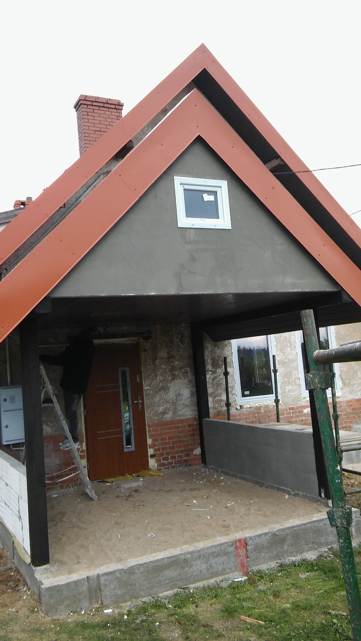 Dom w trakcie budowy z widocznym nowym zadaszeniem nad wejściem, oknem na poddaszu, ceglanym kominem i pracownikiem na drabinie przy kolumnie.